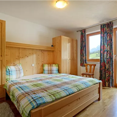 Ein gemütliches Schlafzimmer mit Holzmöbeln und einem großen Fenster. Die Aussicht zeigt eine grüne Landschaft und Berge im Hintergrund.