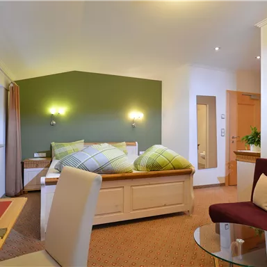 Ein gemütliches Hotelzimmer mit einem Doppelbett, einem roten Sessel und einem kleinen Tisch. Sanfte Farben und Pflanzen sorgen für eine einladende Atmosphäre.