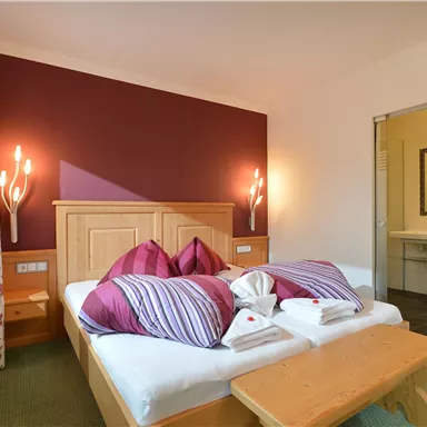 Ein gemütliches Schlafzimmer mit einem Doppelbett, bunten Kissen und Holzverkleidung. Im Hintergrund ist das Badezimmer sichtbar.