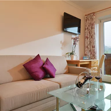 Ein gemütliches Wohnzimmer mit einem beigen Sofa und lila Kissen. Auf dem Tisch stehen eine Karaffe und zwei Weingläser, während große Fenster für Tageslicht sorgen.