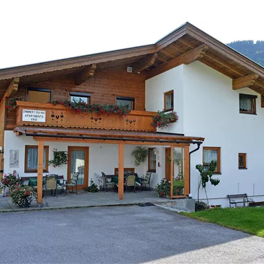 Ein schönes Holzhaus mit einem Balkon und einem begrünten Bereich. Im Hintergrund sind sanfte Hügel zu sehen.