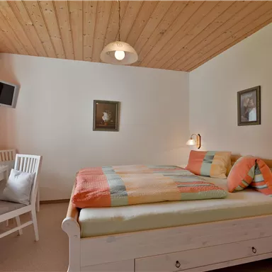 Ein gemütliches Schlafzimmer mit einem Doppelbett und bunten Bettdecken. Der Raum hat einen Schreibtisch und Fenster mit Vorhängen.