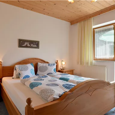 Ein gemütliches Schlafzimmer mit einem Holzbett und bunten Bettwäsche. Es gibt einen großen Fensterblick auf einen Balkon mit Aussicht.