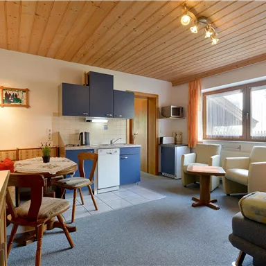 Ein gemütliches Wohnzimmer mit einer modernen Küchenecke und einem Esstisch. Große Fenster sorgen für viel Tageslicht und eine einladende Atmosphäre.