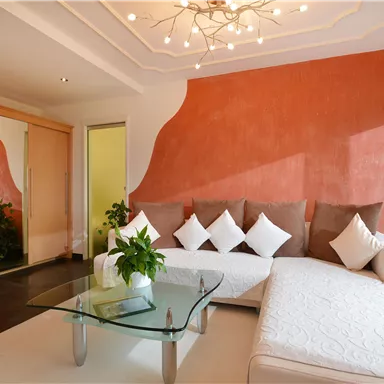 Ein modernes Wohnzimmer mit einer weißen Couch und dekorativen Kissen. An der Wand ist ein warmer Orangeton, und ein eleganter Glastisch ergänzt den Raum.