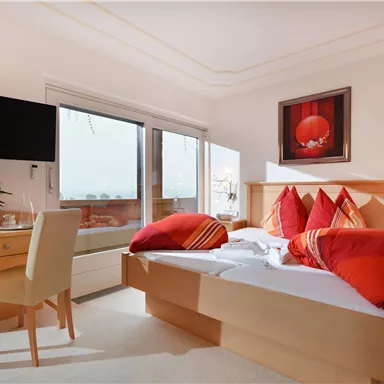 Ein gemütliches Schlafzimmer mit einem großen Bett und bunten Kissen. Es gibt einen Schreibtisch, einen Fernseher und einen Balkon mit Blick nach draußen.