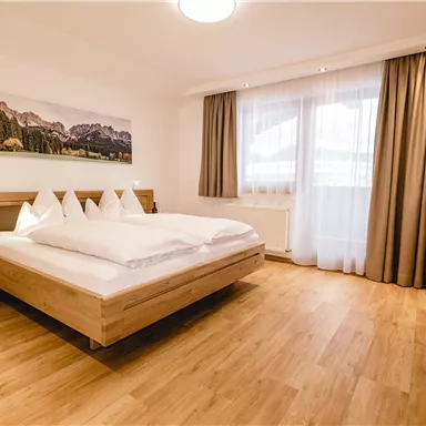 Ein modernes Schlafzimmer mit einem Doppelbett und Holzfußboden. Große Fenster lassen viel Tageslicht herein und sorgen für eine gemütliche Atmosphäre.