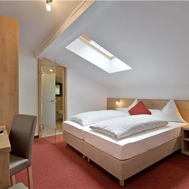 Ein modernes Schlafzimmer mit einem Doppelbett und einem Schreibtisch. Der Raum ist hell und freundlich, mit einem Dachfenster und Holzmöbeln.