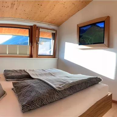 A modern bedroom with a large bed and cozy bedding. A television and a mirror hang on the wall, and light shines through the windows.