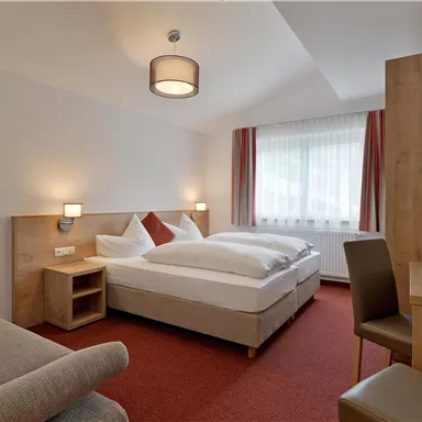 Ein gemütliches Hotelzimmer mit einem Doppelbett und einem Schreibtisch. Die großen Fenster sorgen für viel Tageslicht.