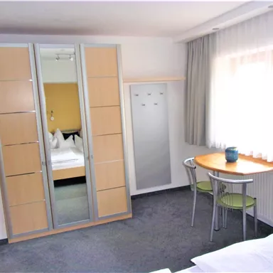 Ein gemütliches Zimmer mit einem Spiegelkleiderschrank und einem Tisch mit Stühlen. Helle Vorhänge lassen viel Licht herein.