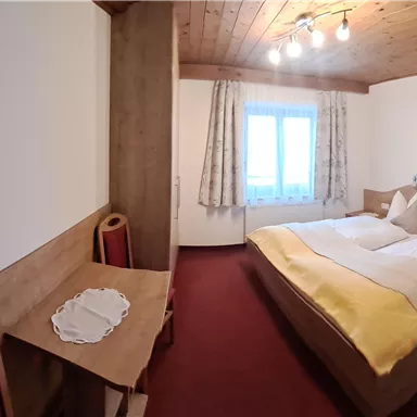 Ein gemütliches Zimmer mit zwei Betten, Holzdecke und Teppichboden. Ein kleiner Tisch und ein Fenster mit Vorhängen vervollständigen die Einrichtung.