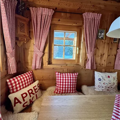 A cozy wooden room with checkered curtains and cushions. A window at the table offers a view outside.