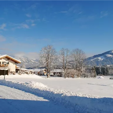 A winter landscape with snow-covered fields and mountains in the background. A cozy house stands in the foreground under a clear blue sky.