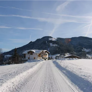Eine verschneite Landschaft mit einem Wohnhaus und einer ruhigen Straße. Im Hintergrund sind sanfte Berge und ein klarer Himmel zu sehen.