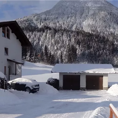 Eine verschneite Landschaft mit einem weißen Haus im Vordergrund. Im Hintergrund erhebt sich ein hoher, schneebedeckter Berg unter einem klaren Himmel.