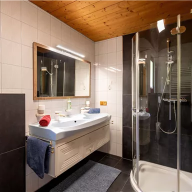 Ein modernes Badezimmer mit einer Dusche und einem großen Spiegel. Die Wände sind hell gefliest, und es gibt ein Waschbecken mit verschiedenen Pflegeprodukten.