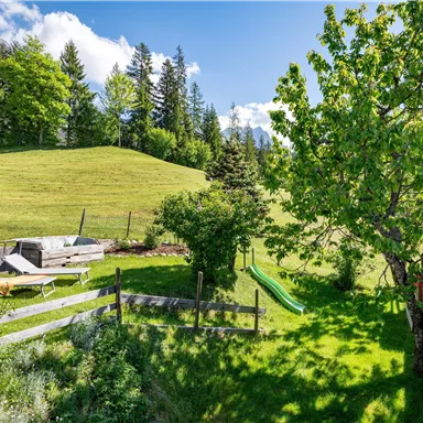 Eine grüne Wiese mit Bäumen und einem Spielplatz. Im Hintergrund sind sanfte Hügel und ein klarer blauer Himmel zu sehen.