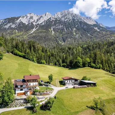 Eine malerische Landschaft mit grünen Wiesen und Bergen im Hintergrund. Im Vordergrund stehen mehrere Häuser, umgeben von Bäumen und einer ruhigen Umgebung.