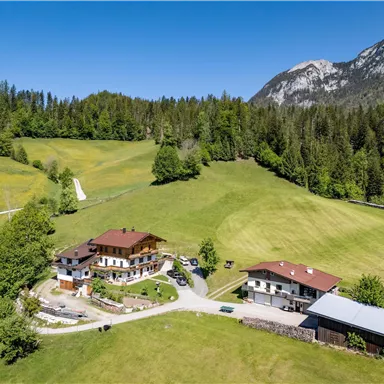Eine malerische Landschaft mit grünen Wiesen und Bergen im Hintergrund. Zwei gut erhaltene Bauernhäuser sind in der Mitte des Bildes zu sehen.