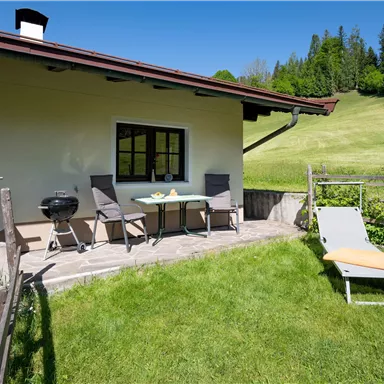 Eine gemütliche Terrasse mit einem Tisch und Stühlen, umgeben von grünem Gras. Im Hintergrund sind sanfte Hügel und ein klarer blauer Himmel sichtbar.
