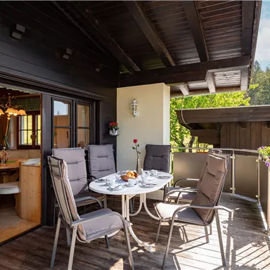 Eine gemütliche Terrasse mit einem Tisch und Stühlen. Im Hintergrund sind Blumen und Bäume zu sehen.