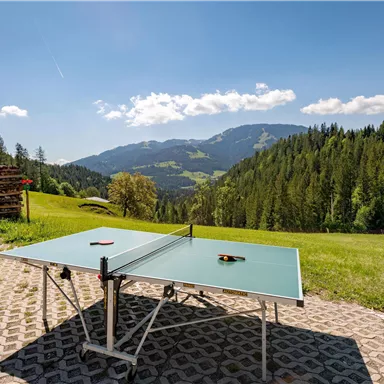 Ein Tischtennistische steht in einer schönen Landschaft mit Bergen und Wäldern im Hintergrund. Der Himmel ist klar und die Sonne scheint.