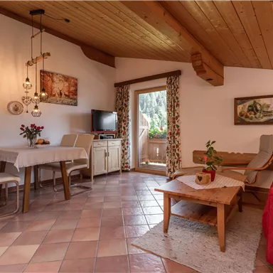 Ein gemütliches Wohnzimmer mit Holzbalkendecke. Es gibt einen Esstisch, eine rote Couch und einen Zugang zu einem Balkon mit Aussicht.