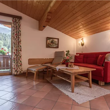 Ein gemütliches Wohnzimmer mit einem roten Sofa und Holzbalkendecke. Durch das Fenster sieht man einen Blick auf die Berge und frische Blumen.