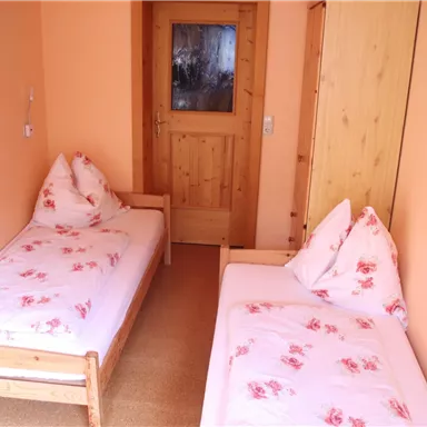 Ein einfaches Zimmer mit zwei Einzelbetten und rosafarbenen Bettwäsche. Die Wände sind orange, und eine Holzoberfläche ergänzt die Einrichtung.