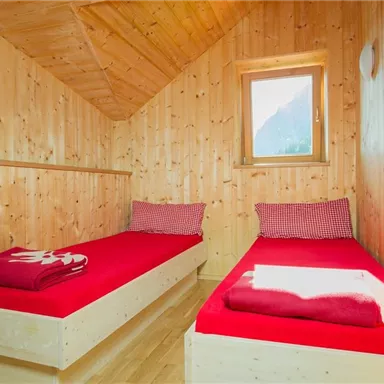 Ein gemütliches Zimmer mit zwei Einzelbetten und roten Bettdecken. Die Wände sind aus Holz, und es gibt ein Fenster mit Blick auf die Natur.