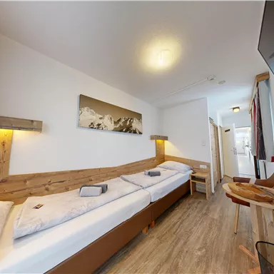 A modern room with two single beds, wooden details, and a television. The walls are light, and there is a small table as well as a chair.