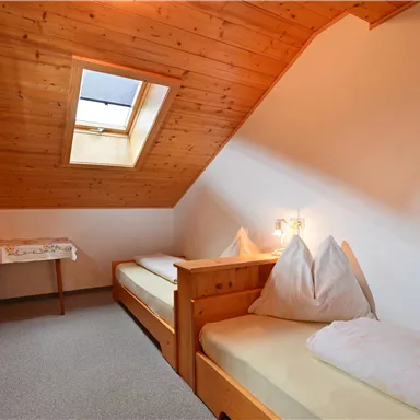 Ein gemütliches Zimmer im Dachgeschoss mit schrägem Holzdecken. Es gibt zwei Einzelbetten und einen kleinen Tisch.