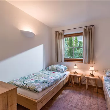 A bright bedroom with two single beds and cozy bedding. By the window are two nightstands with lamps and a wooden wardrobe is in the room.