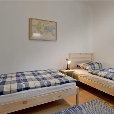 Ein einfaches Zimmer mit zwei Einzelbetten und karierten Bettdecken. An der Wand hängt ein Bild und ein Fenster lässt natürliches Licht hereinscheinen.