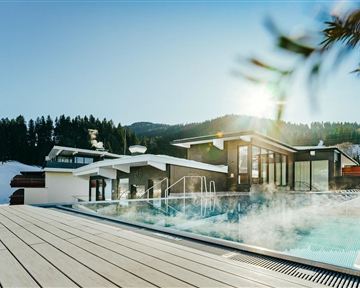 Ein moderner Außenpool umgeben von schneebedeckten Bergen. Die Sonne strahlt und sorgt für eine entspannende Atmosphäre.