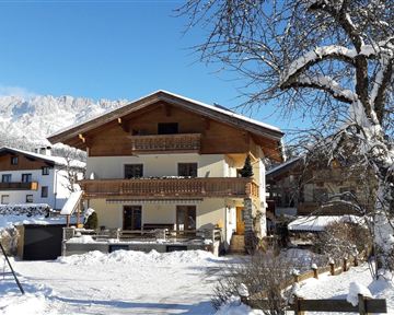 Ein gemütliches Haus im Schnee mit einem klaren blauen Himmel. Umgeben von Bergen und anderen Gebäuden, bietet es eine idyllische Winterlandschaft.