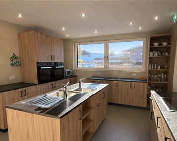 Eine moderne Küche mit Holzschränken und schlichten grauen Arbeitsflächen. Große Fenster sorgen für viel Tageslicht.