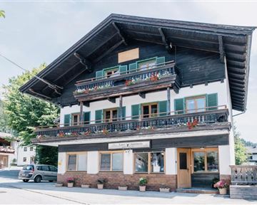 Ein traditionelles, mehrstöckiges Holzhaus mit Balkonen und bunten Blumenkästen. Es liegt in einer ländlichen Umgebung mit grünen Bäumen und einem klaren Himmel.