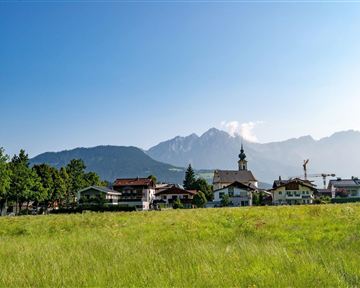 Eine malerische Landschaft mit einem Dorf im Vordergrund und majestätischen Bergen im Hintergrund. Der Himmel ist klar und die Wiese grün und einladend.