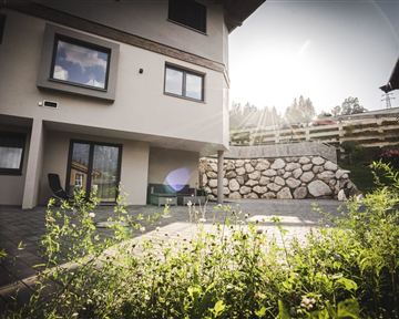 Ein modernes Haus mit einem sonnigen Außenbereich und einem geschmackvoll gestalteten Garten. Die Wände sind hell und die Fenster bieten einen Blick in die Umgebung.