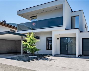 Ein modernes, zweistöckiges Haus mit glatter Fassade und großen Fenstern. Vor dem Haus befindet sich ein kleiner Garten mit einem Baum.