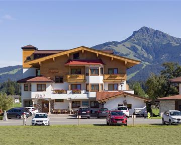 A charming hotel with wooden elements, surrounded by green meadows and mountains. The sky is clear and sunny, creating an inviting atmosphere.