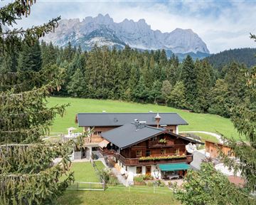 A picturesque mountain cabin surrounded by dense forests and meadows. In the background, majestic mountains rise under a clear sky.