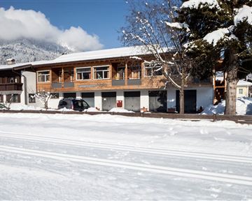 A winter landscape with a wooden building and snow-covered ground. In the background, mountains are visible under a blue sky.