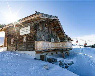 Ein gemütliches Holzhaus in einer verschneiten Landschaft. Im Hintergrund sind Skilifte zu sehen, und die Sonne scheint hell am Himmel.