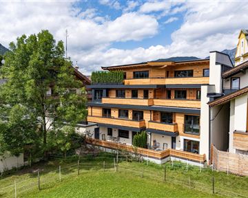 A modern building with wooden cladding and several balconies. In the foreground, there is a green lawn with some trees visible.