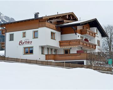 Ein gemütliches Haus im alpinen Stil, umgeben von Schnee. Im Hintergrund sind Berge sichtbar, was eine malerische Winterlandschaft schafft.
