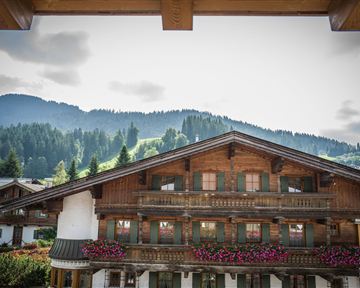 Ein traditionelles Holzhaus mit bunten Blumenbalkonen und Blick auf die Berge. Der Himmel ist bewölkt und die Umgebung ist grün und hügelig.