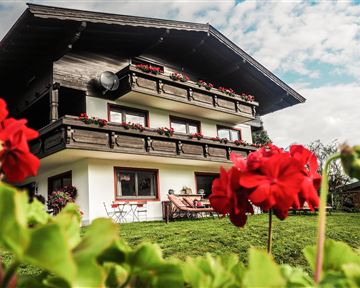 Ein charmantes Haus mit Holzverkleidung und mehreren Balkonen. Vor dem Haus blühen rote Blumen in einer grünen Wiese.